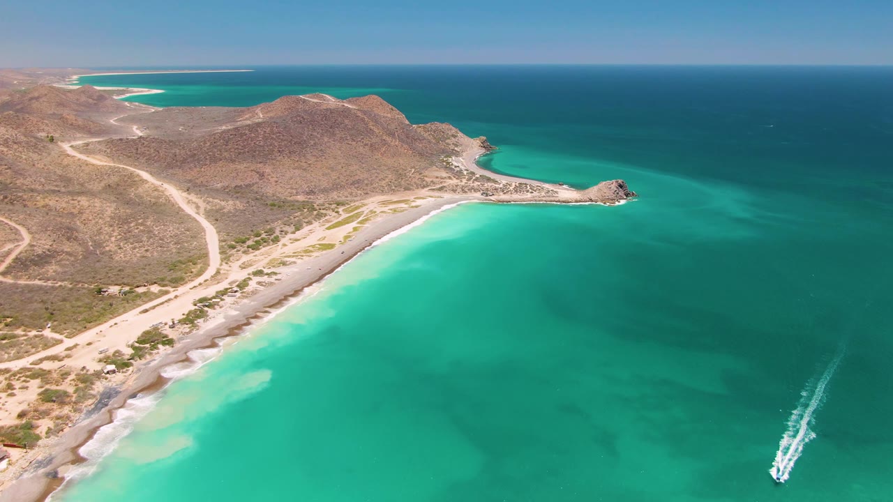 Aerial frame of Cabo Pulmo coastline with the boat slightly farther along the lower right water, its wake bright against turquoise shallows, with beach, hills, roads, and no visible people.