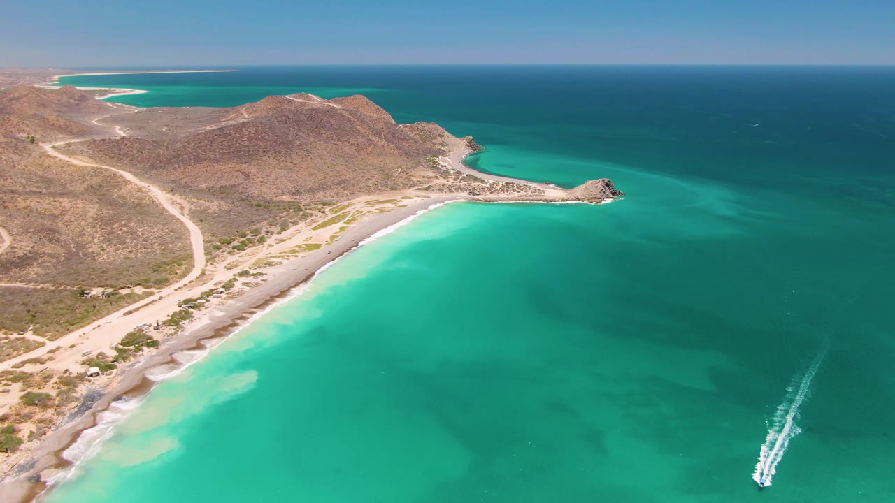 Wide aerial view over Cabo Pulmo with the boat wake near the lower right edge, clear turquoise water filling the foreground, dry hills and dirt roads along shore, and no visible people.