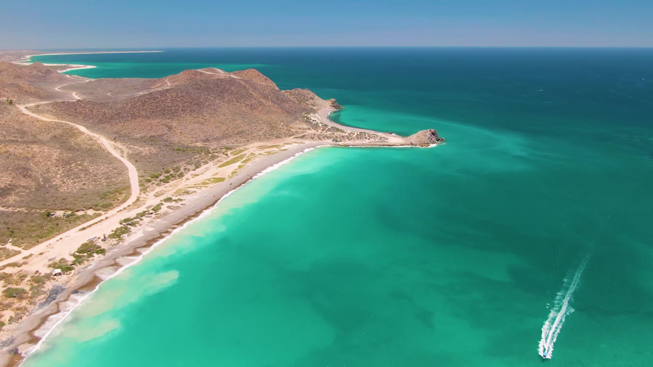 Cabo Pulmo aerial frame with more open water in the foreground, the boat wake near the lower right border, a pale beach curving below dry hills, and no visible people.