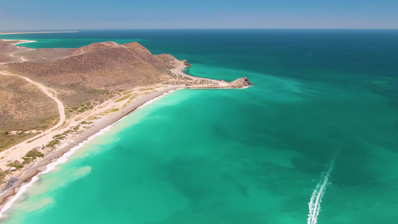 Aerial frame over Cabo Pulmo showing turquoise shallows below, a curved sandy beach and dirt road at left, arid hills behind, deep blue ocean on the horizon, and no visible people.