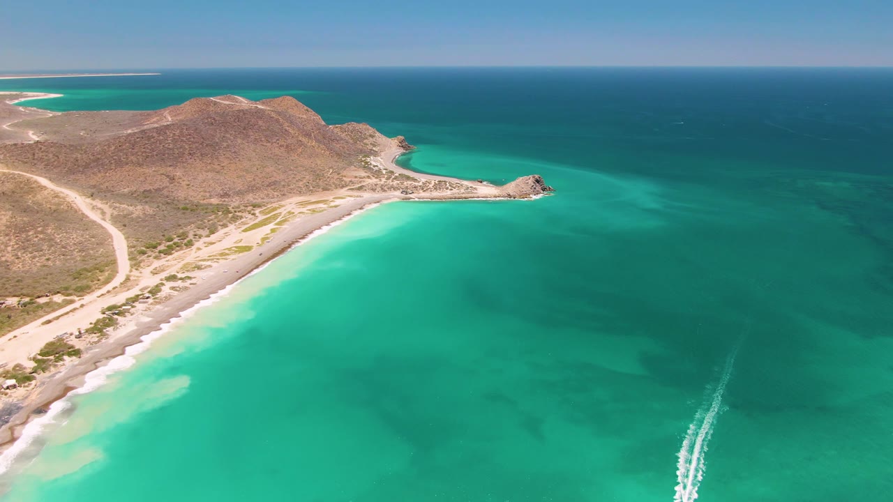 Drone view of a curved beach beside turquoise water with a boat wake near the lower right.