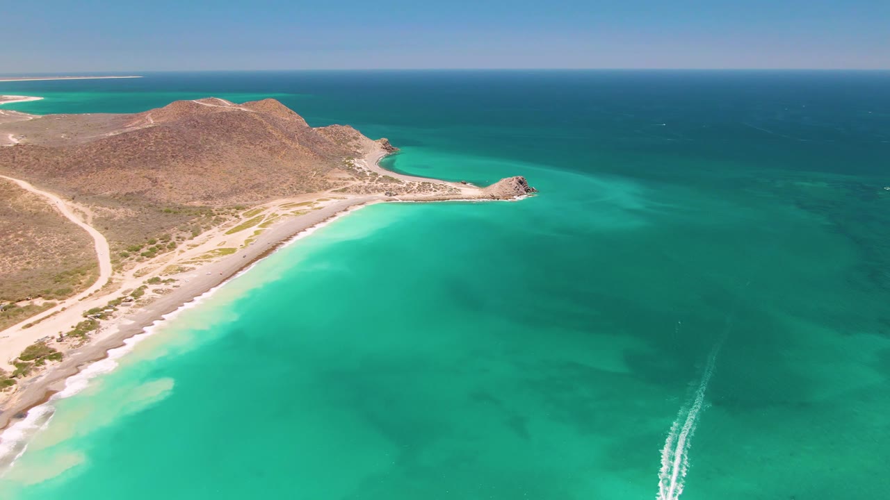 Aerial frame over Cabo Pulmo with the boat wake farther up through the turquoise water, a rocky point and sandy beach along the left side, dry hills in back, and no visible people.