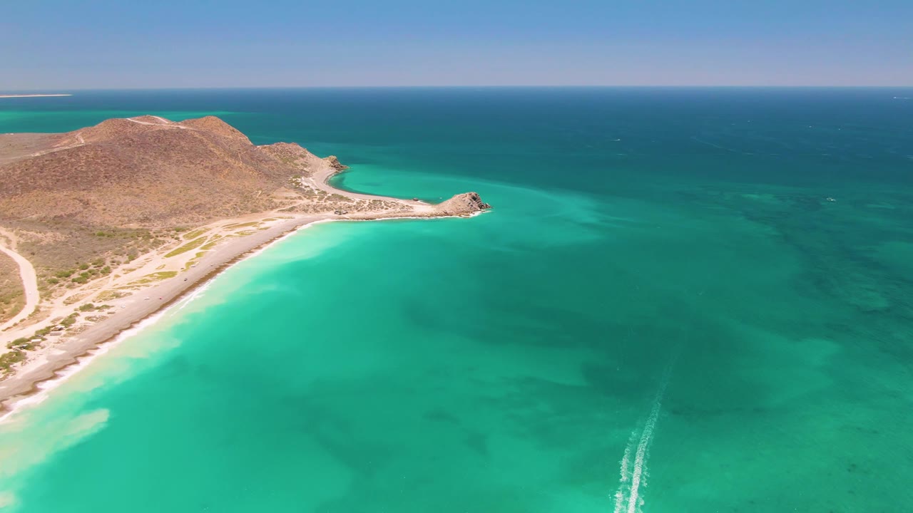 At 0:00, an aerial view over Cabo Pulmo shows turquoise water, a pale curved beach, a dry hill, a coastal road at the far left, a faint boat wake, and no visible people.