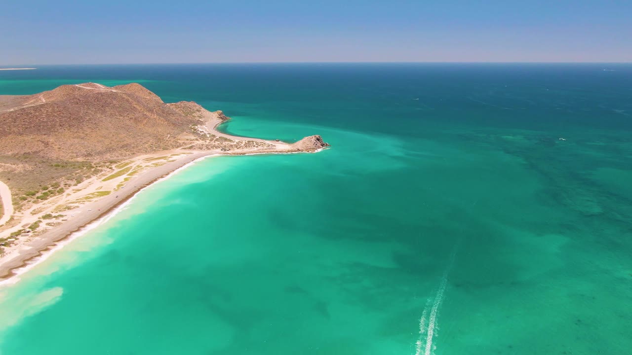At 0:01, the Cabo Pulmo aerial holds on the beach curve and brown hillside while the boat wake appears lower in the turquoise water, with no visible people.