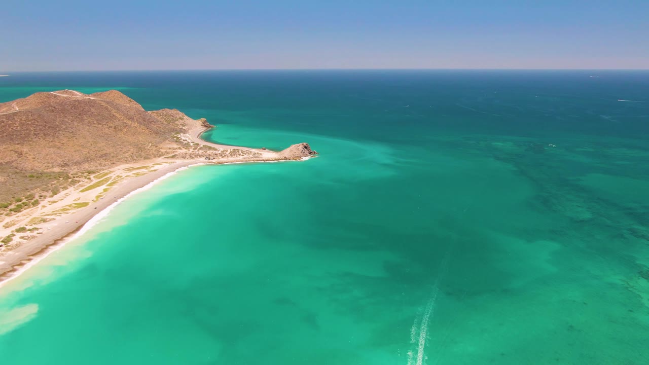 At 0:03, the aerial frame shows more of the road and beach edge on the left, with the dry Cabo Pulmo hill above and clear turquoise water across the frame, with no visible people.