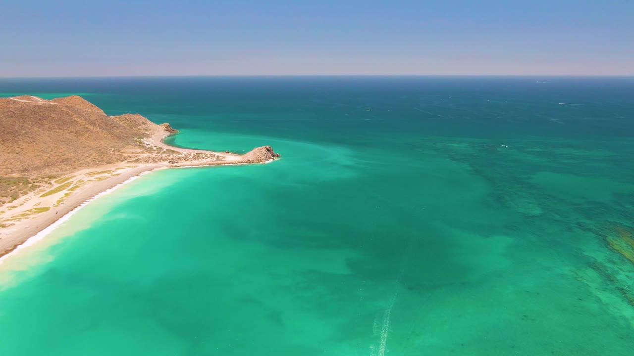 Aerial frame over Cabo Pulmo showing turquoise water, pale sand, a tan rocky headland on the left, a level horizon, and no visible people.