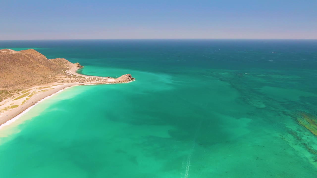 Cabo Pulmo aerial frame with the curved sandy beach and rocky point slightly farther left while clear turquoise ocean stretches toward the horizon, with no visible people.