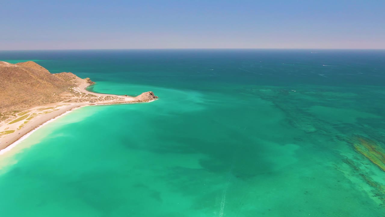 Wide Cabo Pulmo frame showing the rocky headland and beach along the left edge, shallow turquoise water in the foreground, darker reef-like seabed marks, and no visible people.