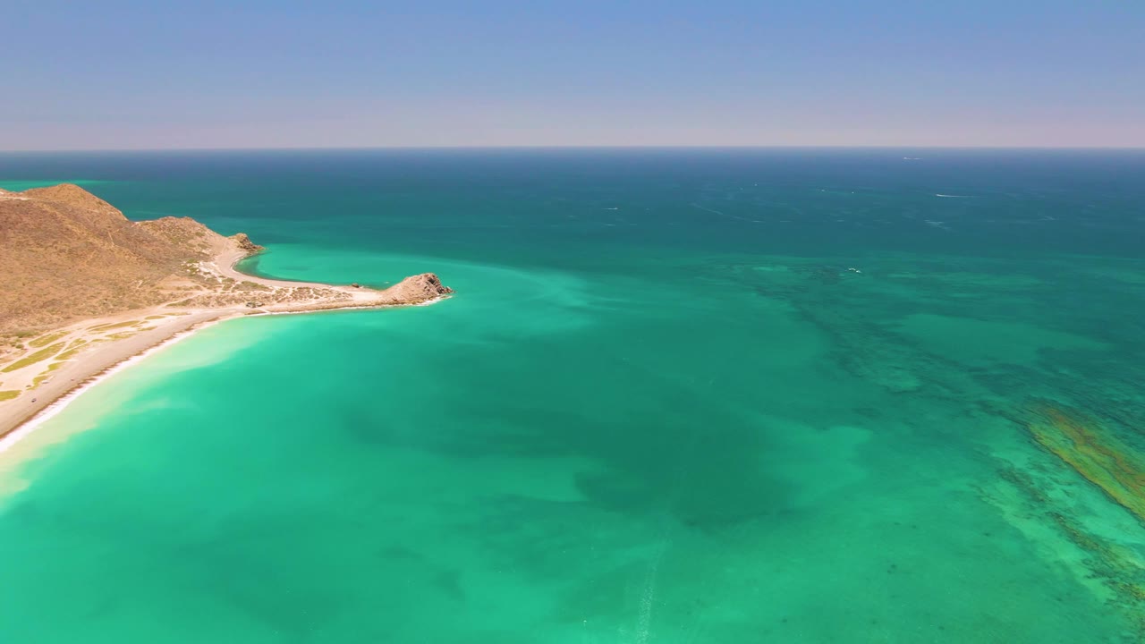 Aerial coastal frame with beach and headland near the left edge and open water to the right.