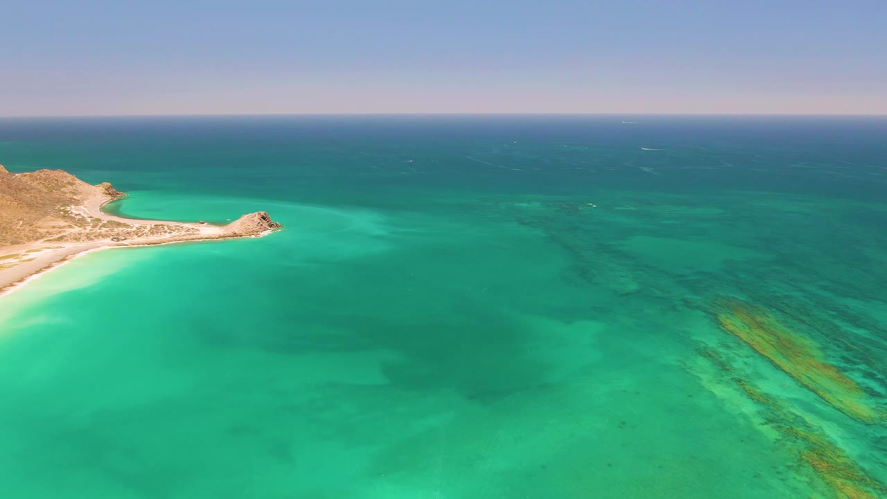 Second frame of Cabo Pulmo coastline with the sandy curve and rocky point slightly farther inside the left side of the frame, broad turquoise water below, and no visible people.