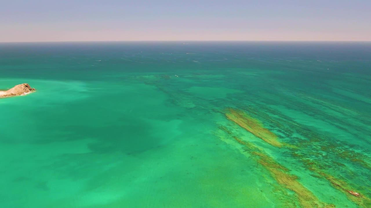 Aerial frame from Cabo Pulmo, Mexico, showing the island slightly right of center, green shallow water in the foreground, darker blue water beyond, reef bands, and no visible people.