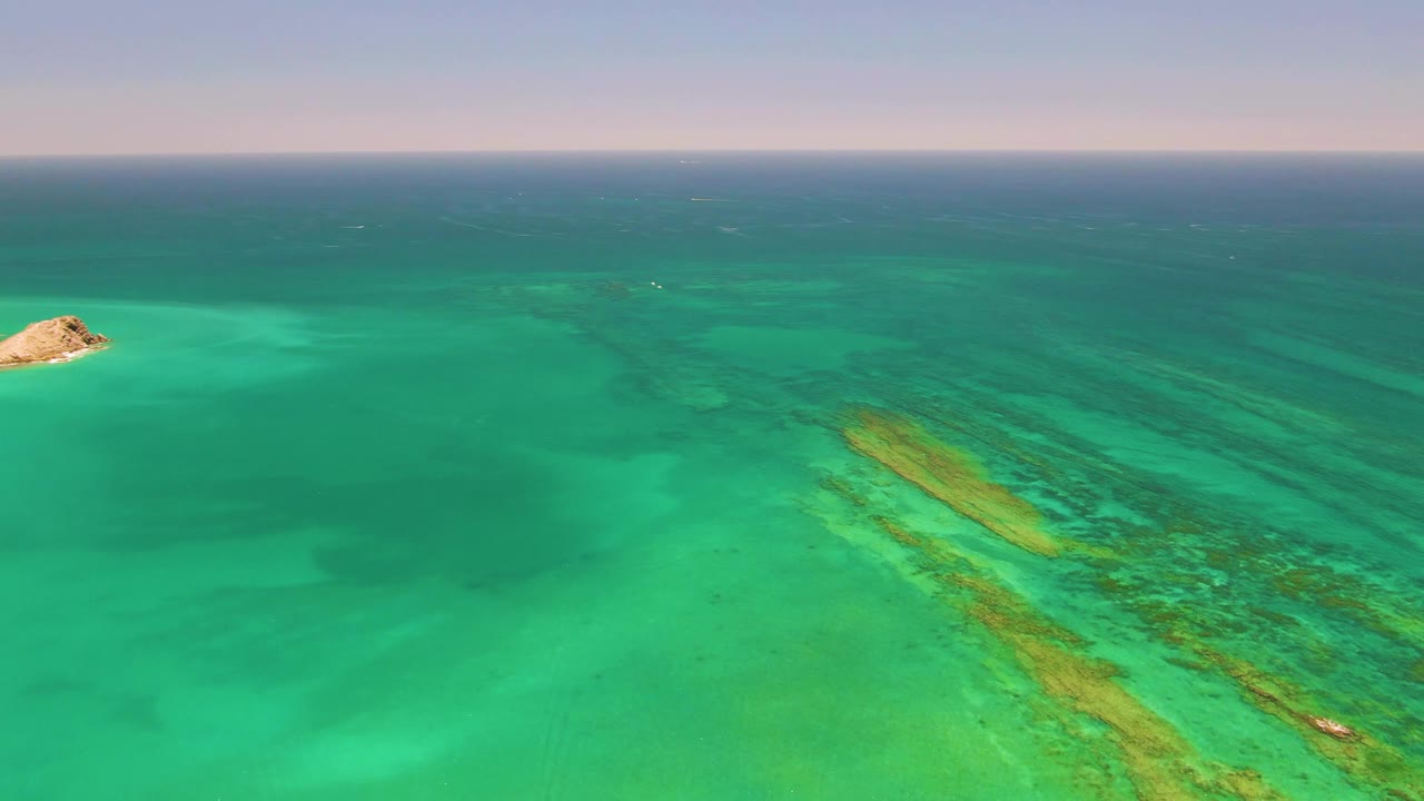 Drone-like aerial view in Cabo Pulmo, Mexico, with the rocky island near the upper center, long yellow-green reef streaks in the lower right water, and no visible people.