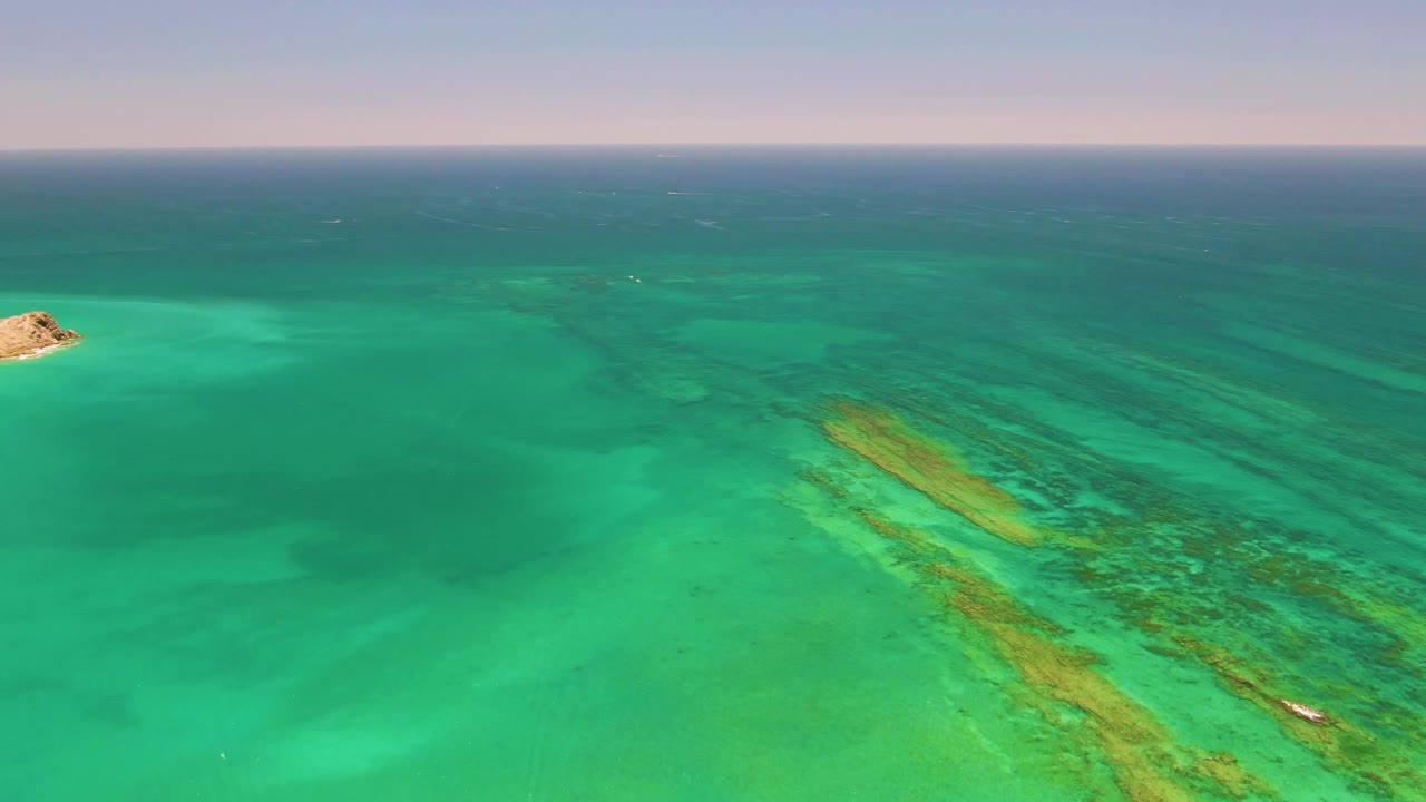 Aerial Cabo Pulmo frame with the island partly at the left edge, coral reef bands crossing the lower right, a small boat near the reef, and no visible people.