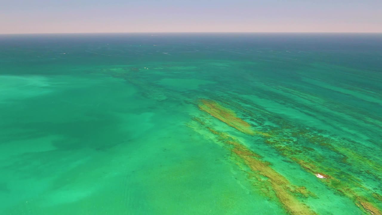 Aerial frame over Cabo Pulmo showing the reef band shifted slightly across the shallow green water, the small white boat beside it, a level sea horizon, and no visible people.