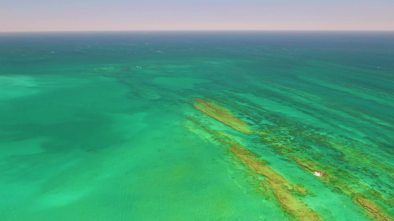 Cabo Pulmo aerial frame with the diagonal reef occupying the lower half of the image, a small white boat near the reef's brighter patch, soft sky at the horizon, and no visible people.