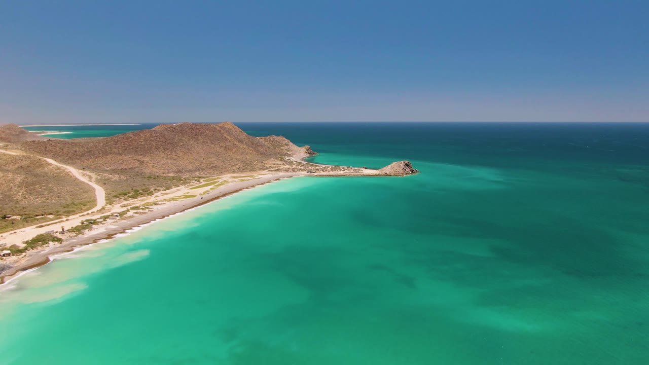 Opening aerial frame over Cabo Pulmo, Mexico, with turquoise water in front, a sandy shoreline and road at left, dry hills beyond, and no visible people.