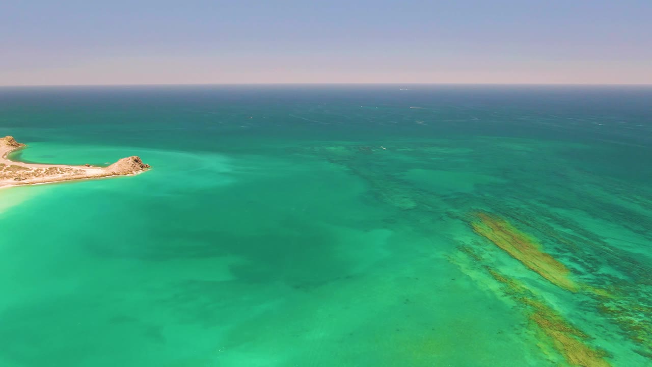 Opening aerial frame over Cabo Pulmo, Mexico, with a small sandy island at the far left, turquoise water across the foreground, darker ocean at the horizon, and no visible people.