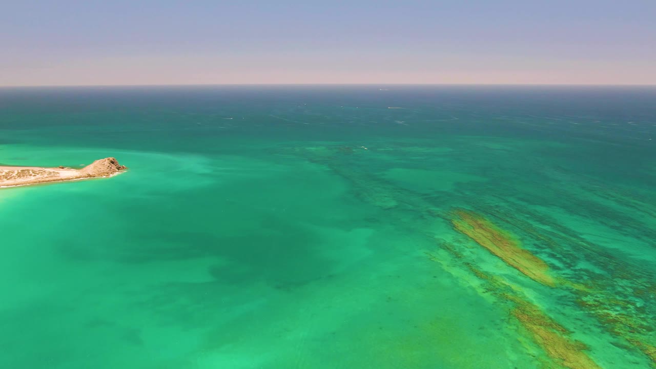 Cabo Pulmo aerial frame with the island reduced near the left side, clear green-blue water filling most of the image, reef-toned streaks on the right, and no visible people.