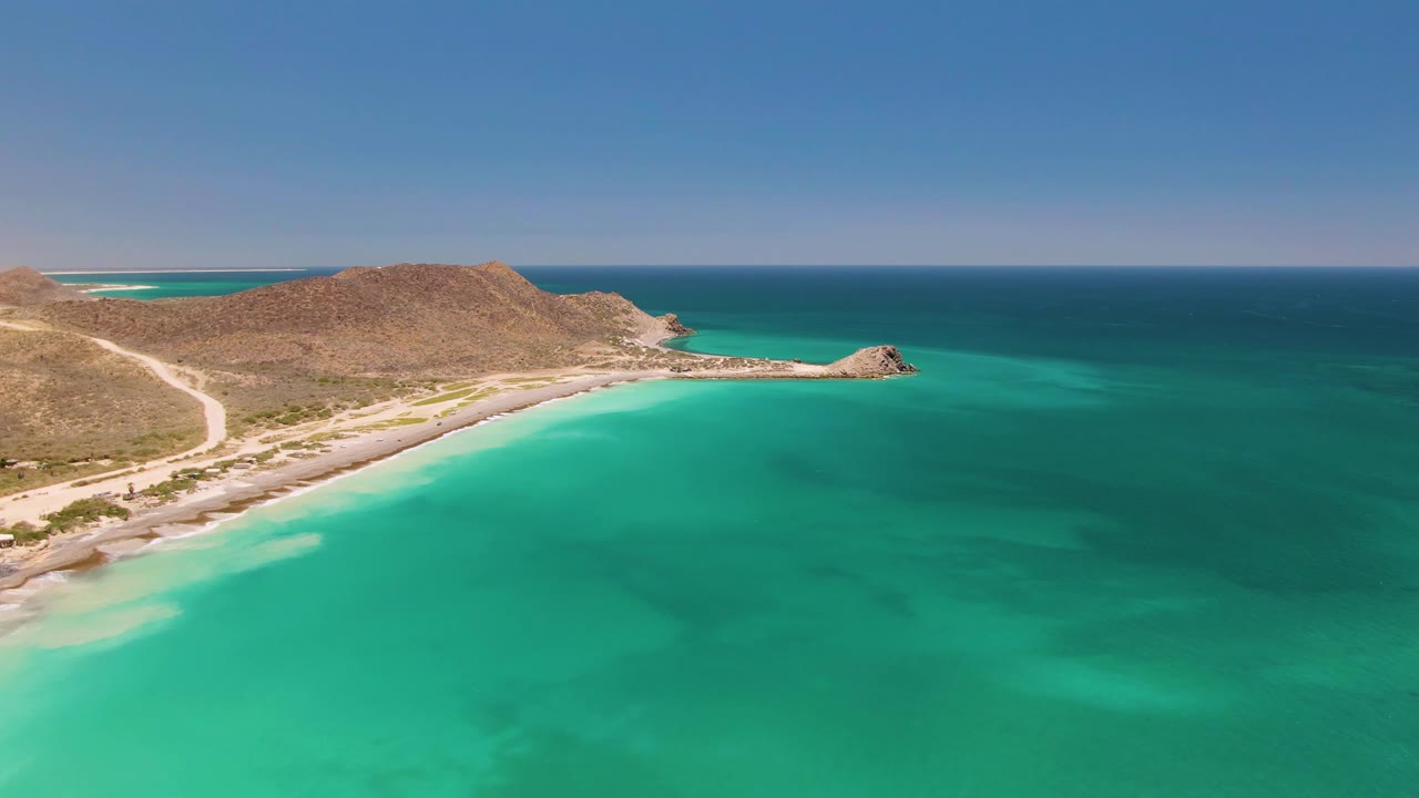 Opening aerial frame over Cabo Pulmo, Mexico, showing turquoise water in the foreground, a sandy beach, a pale coastal road, dry hills, and no visible people.