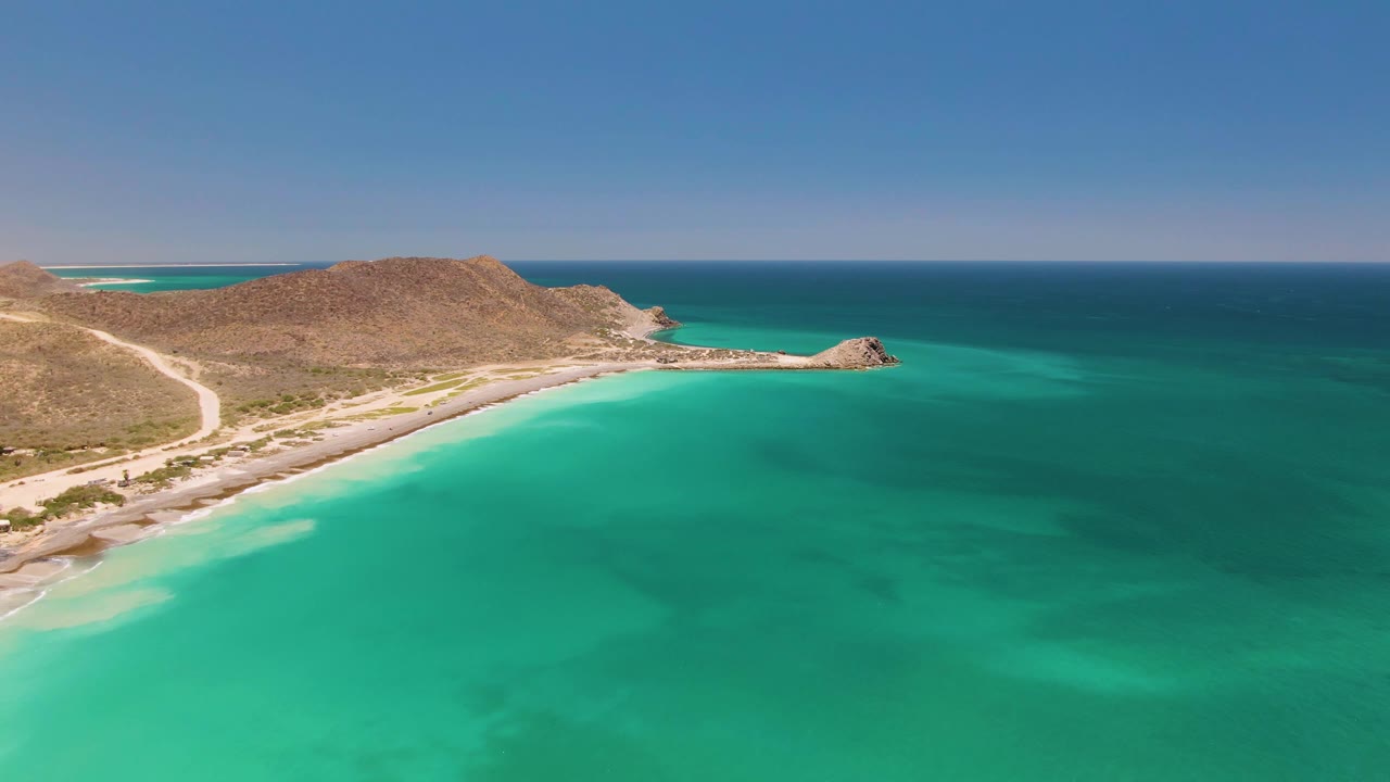 Aerial frame at 0:02 in Cabo Pulmo showing a broad turquoise sea foreground, sandy shore, a road beside arid hills, a rocky point on the coast, and no visible people.