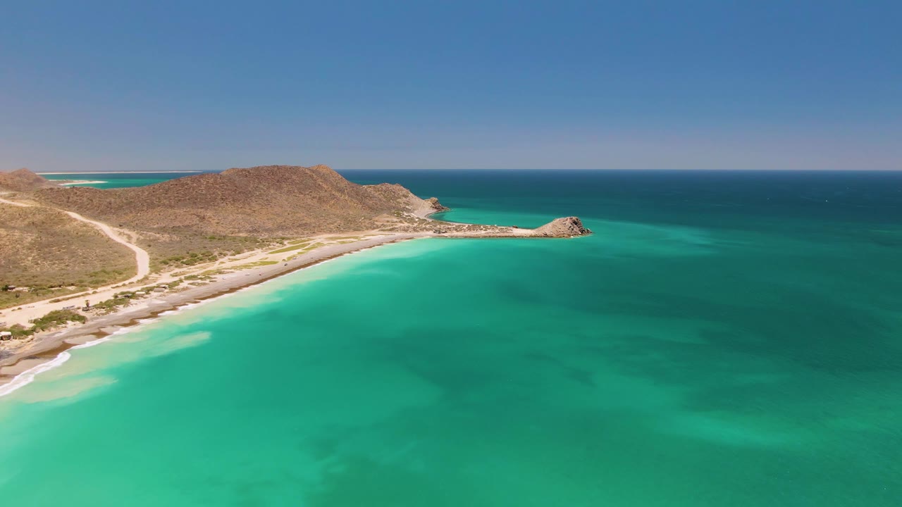 Opening aerial frame over Cabo Pulmo, Baja California Sur, with turquoise water in the foreground, a sandy curved beach, rugged brown hills, and no visible people.