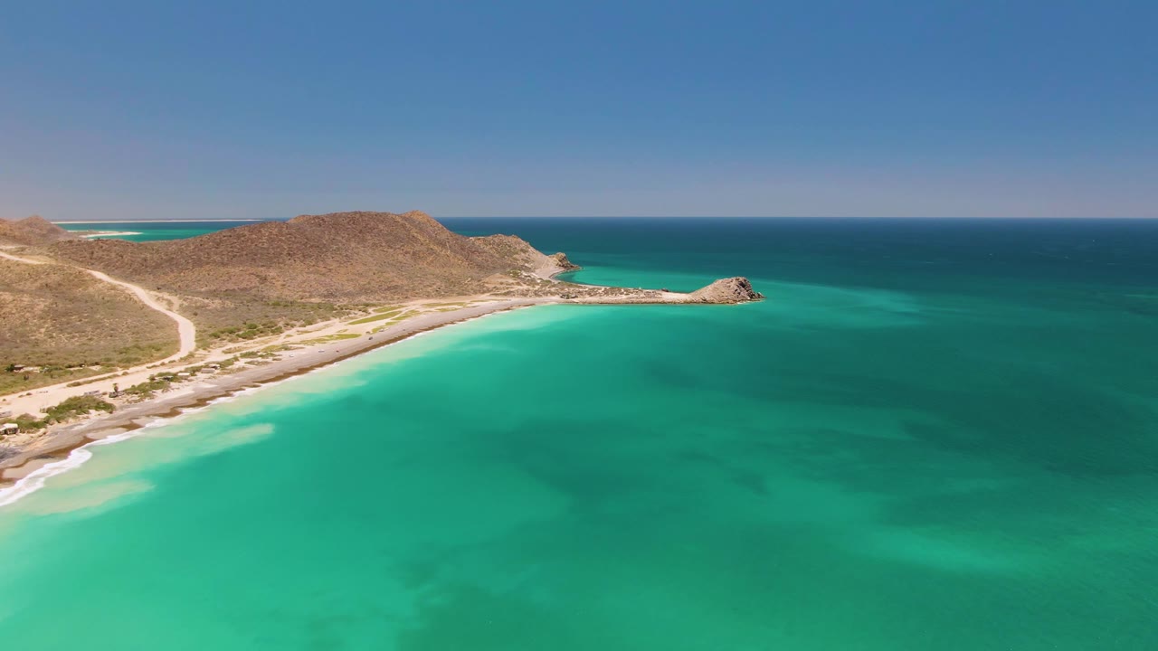Aerial view in Cabo Pulmo showing the sandy shoreline and dirt road along dry hills, with turquoise ocean filling most of the frame and no visible people.