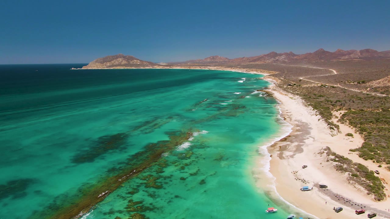 Aerial frame over Cabo Pulmo showing turquoise shallows in front, a pale sandy shore with small boats to the right, distant desert mountains, clear blue sky, and no visible people.