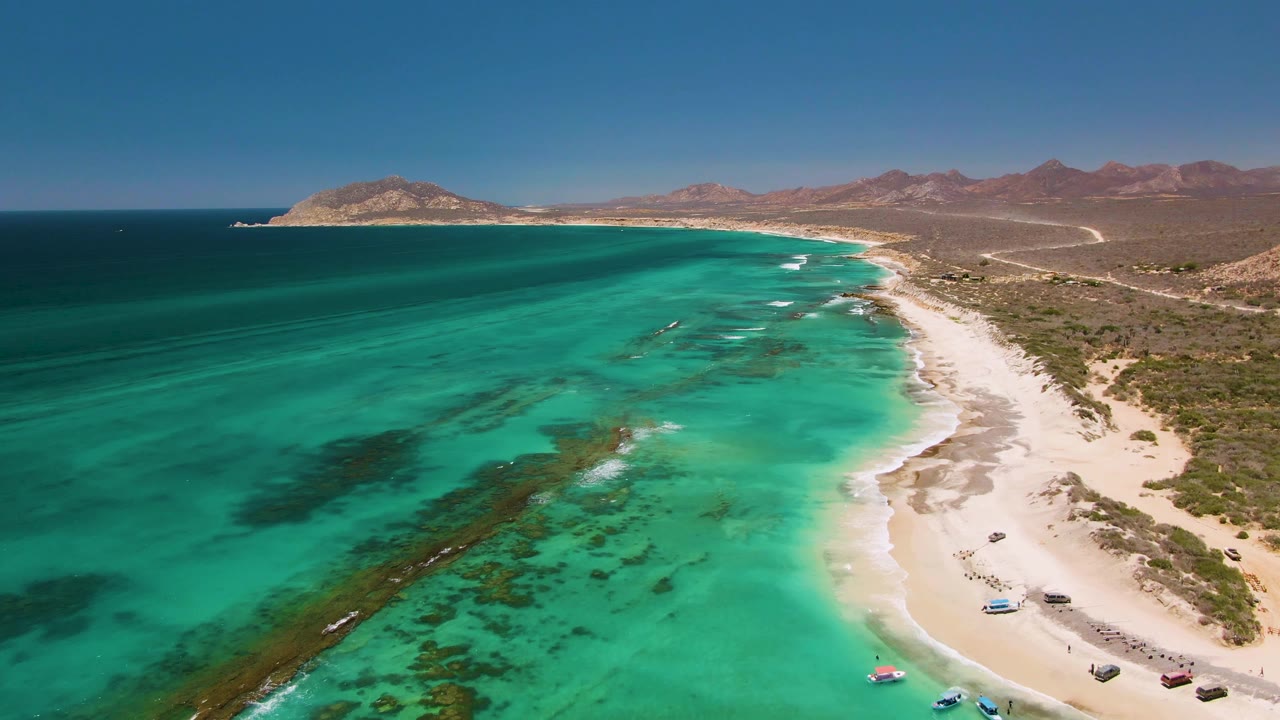 Turquoise coastal water beside a sandy shore with boats and mountains beyond.