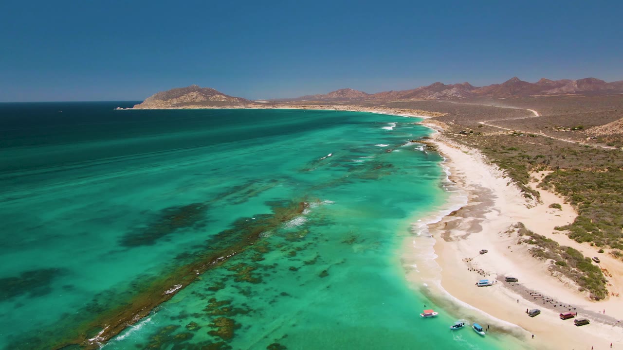 High aerial frame in Cabo Pulmo showing a curved sandy beach on the right, bright turquoise water on the left, several small boats near shore, hazy desert mountains, and no visible people.