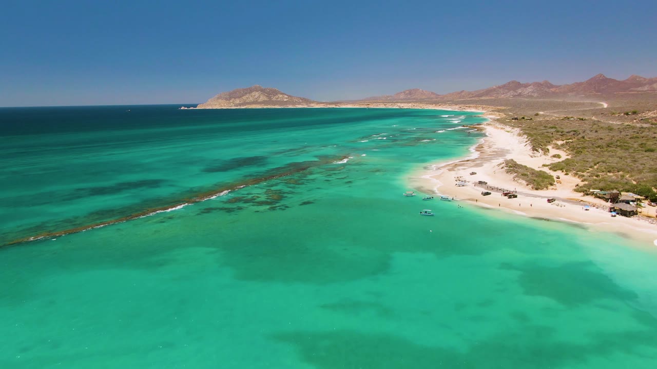 Aerial view of turquoise ocean beside a sandy beach and distant hills.