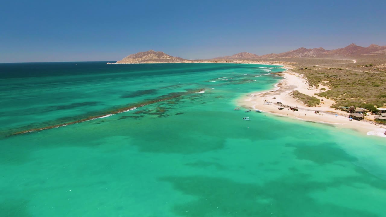 Aerial view over Cabo Pulmo showing a broad turquoise bay, darker underwater patches, a pale beach curving along the right side, low hills beyond, and no visible people.