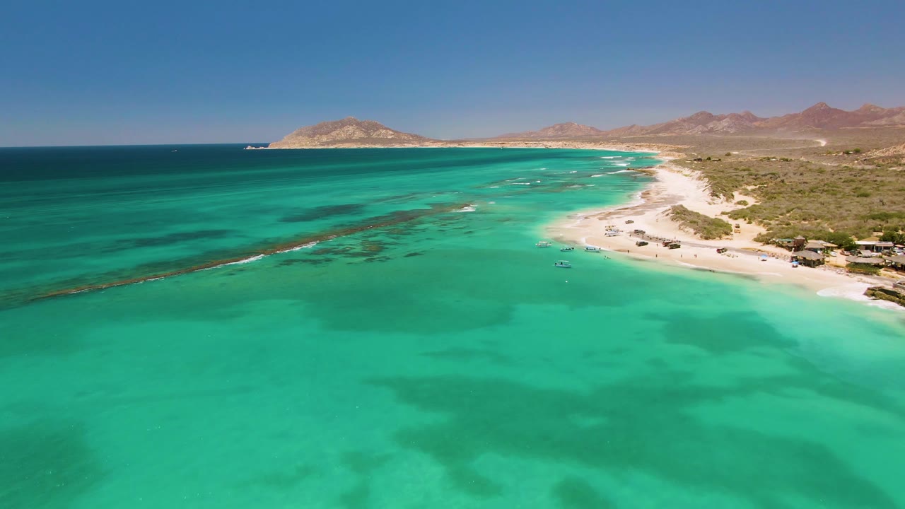 Cabo Pulmo coastline from above with open turquoise water in front, a sandy beach with small structures and vehicles on the right, brown mountain ridges inland, and no visible people.