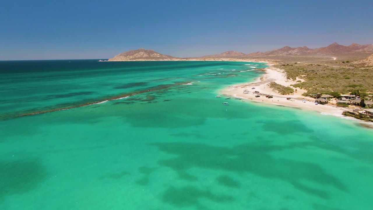Later aerial frame over Cabo Pulmo with more foreground water visible, dark underwater patches near the beach, small structures along the sand, dry hills inland, and no visible people.