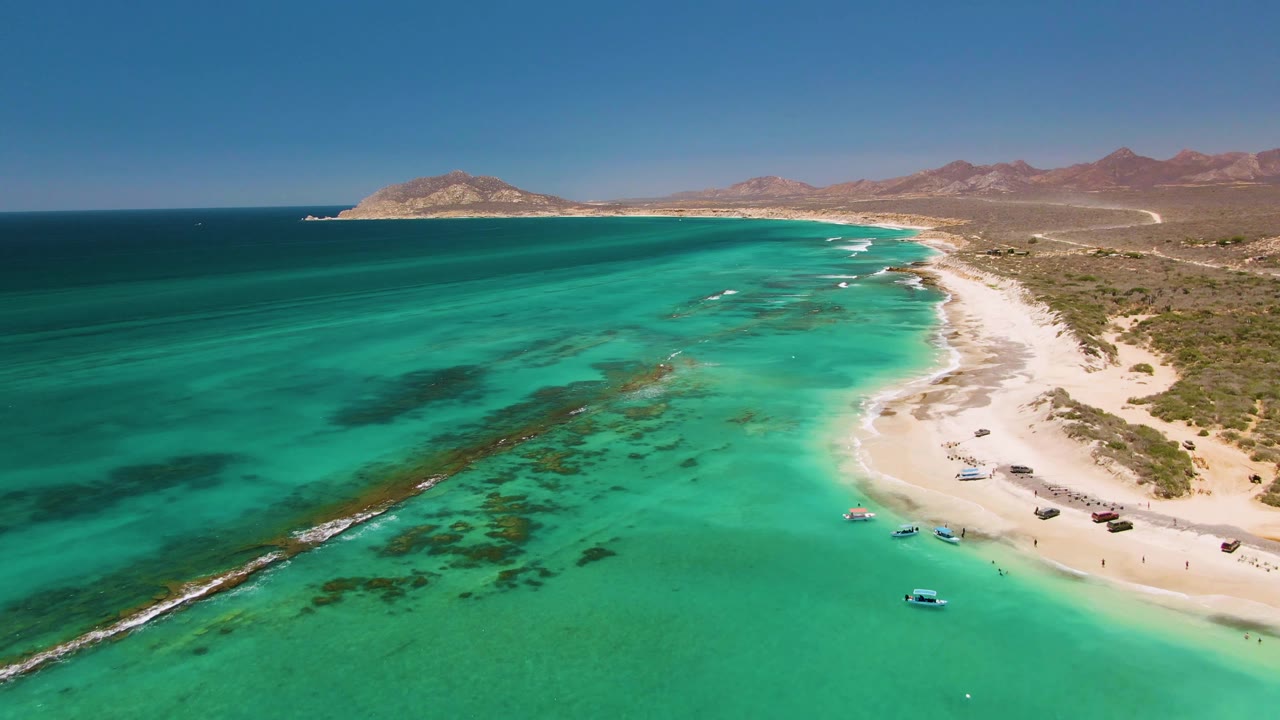 Aerial frame over Cabo Pulmo showing turquoise shallows, a sandy beach with small boats near shore, darker underwater rock lines, arid hills, blue sky, and no visible people.