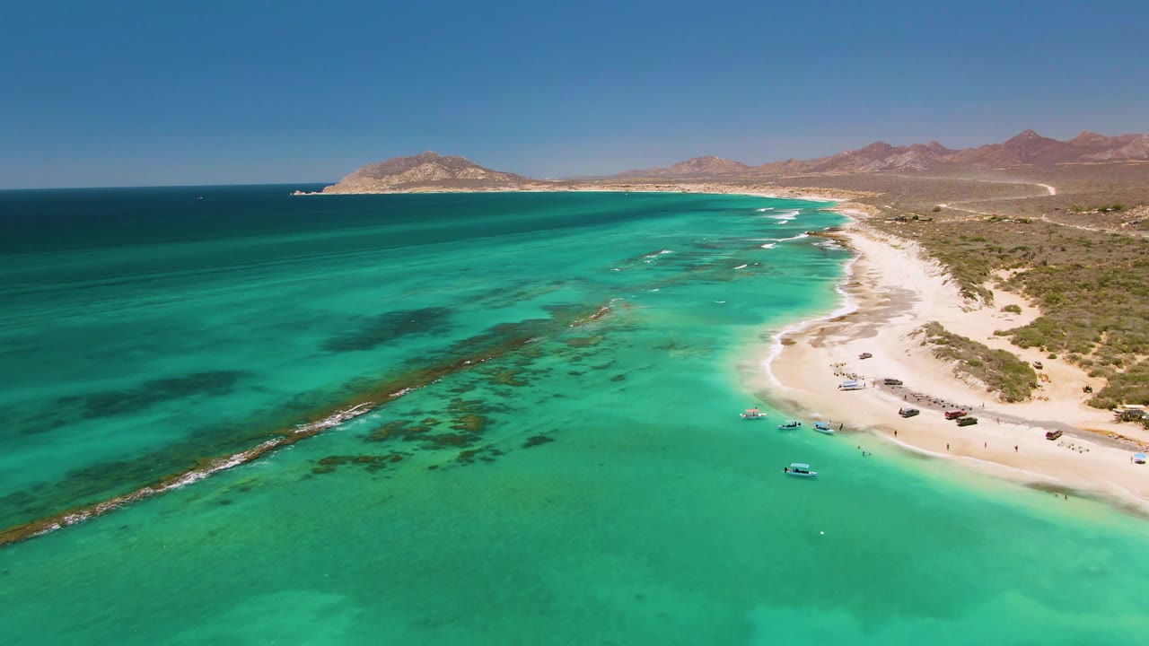 Cabo Pulmo aerial frame with clear aqua water filling most of the view, boats near the right-side beach, thin white surf, distant brown mountains, and no visible people.