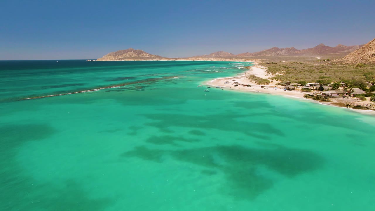 Opening aerial frame over clear turquoise water at Cabo Pulmo, Mexico, with a sandy shoreline, low buildings, arid hills, distant mountains, and no visible people.