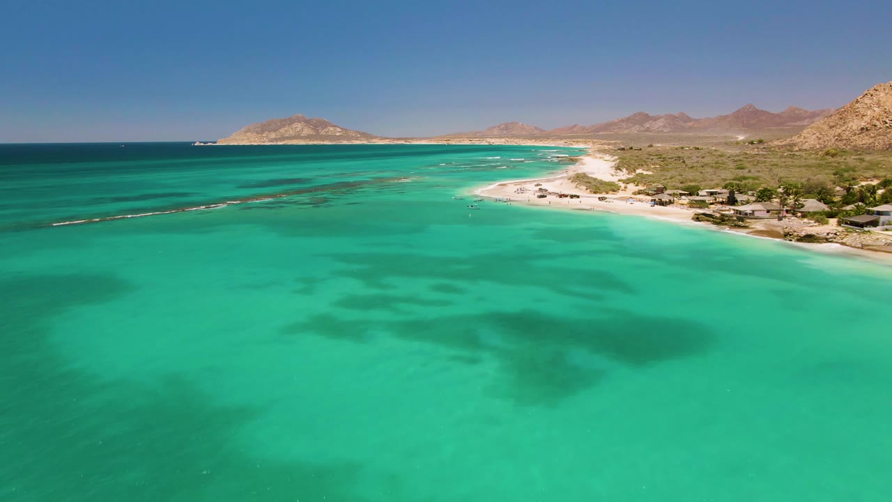 Wide aerial coastline with clear water, sandy beach, and desert hills.