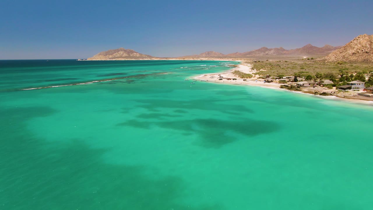 Cabo Pulmo aerial frame with turquoise water occupying most of the view, darker seabed areas below, a sandy beach to the right, small buildings near shore, mountains, and no visible people.