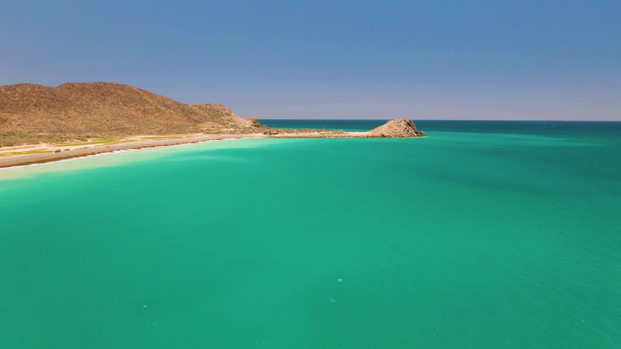 Wide aerial view of turquoise water, pale sand, dry hills, and a small rocky outcrop at Cabo Pulmo, Mexico, with no visible people.