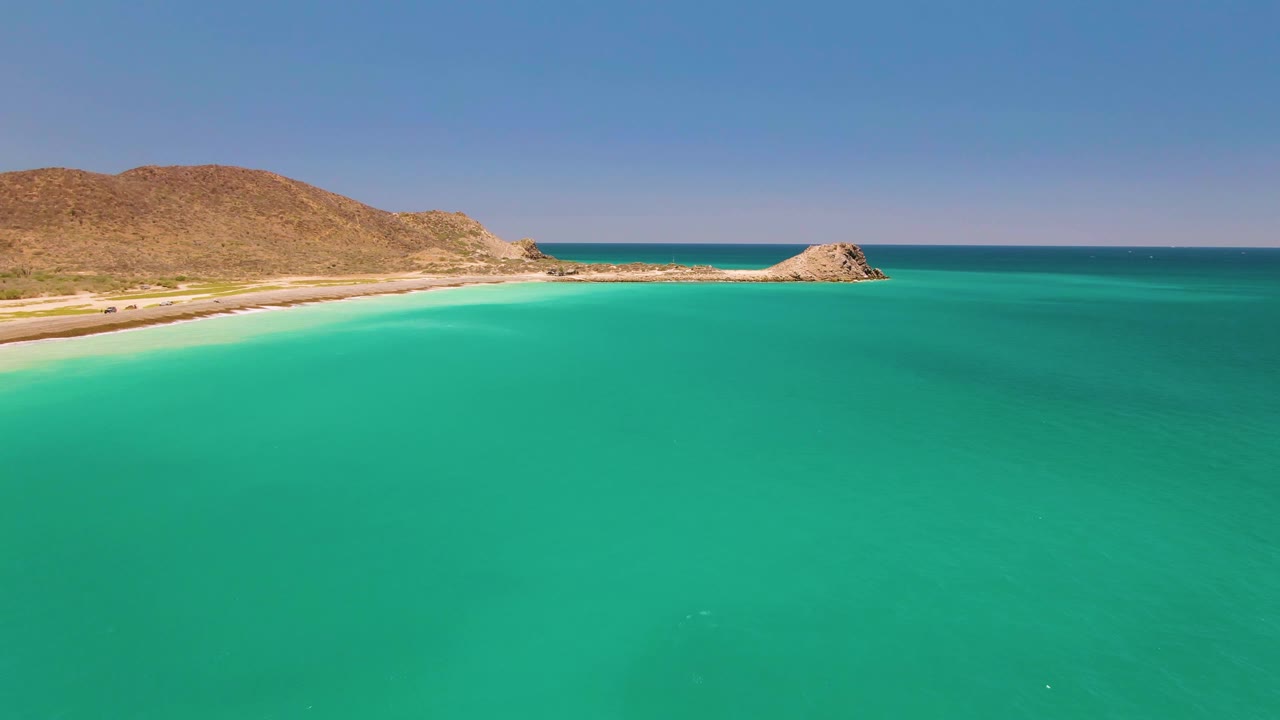 Steady aerial view over calm turquoise water at Cabo Pulmo, with the sandy shoreline and dry hills running across the upper frame and no visible people.