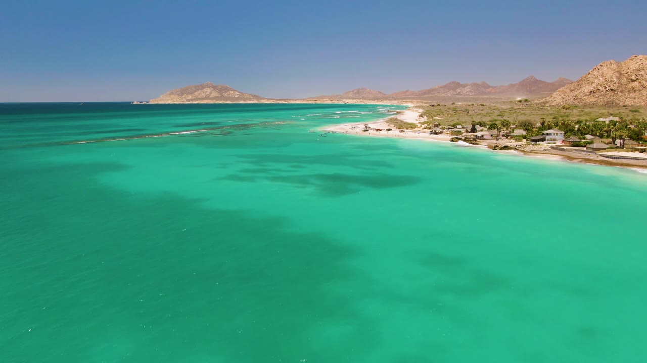 Wide aerial frame over turquoise water at Cabo Pulmo, Mexico, with a sandy shore curving below rocky hills and small shoreline structures visible at the right.