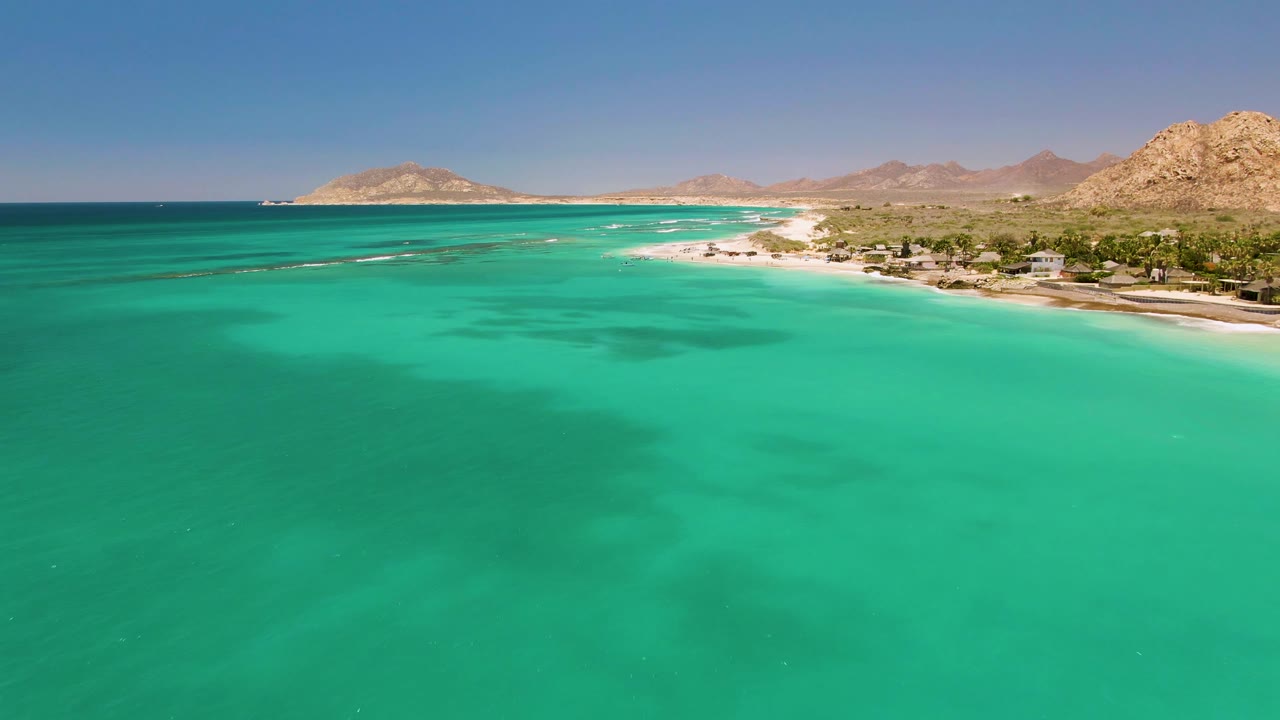 The frame holds a high aerial angle over Cabo Pulmo, Mexico, with clear green-blue water in the foreground, thin surf lines near shore, and tan rocky hills behind the beach.