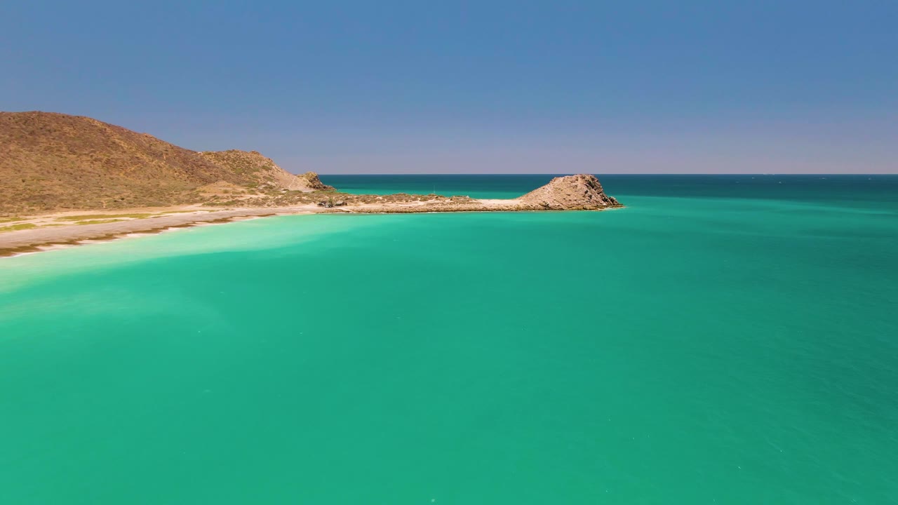 Cabo Pulmo shoreline seen from above with broad turquoise water in the foreground, a pale beach curve at left, rocky shoreline near center, dry hills in back, and no visible people.