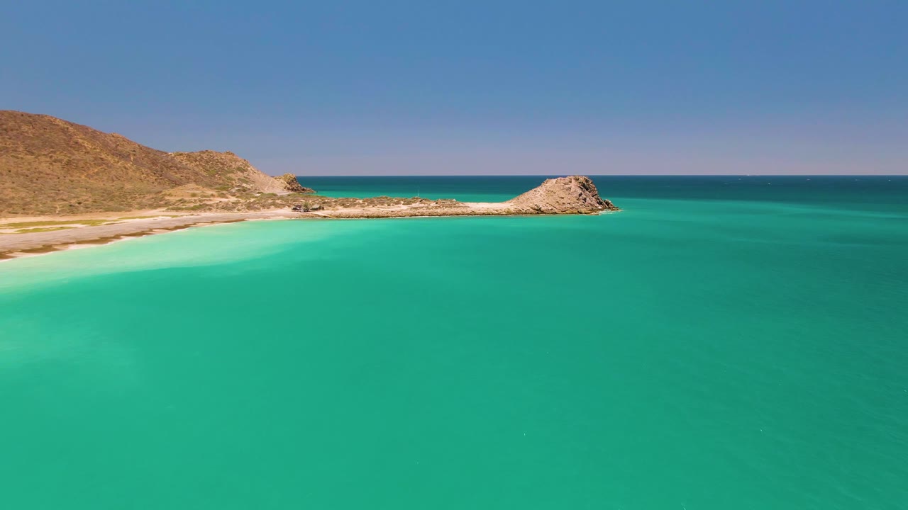 Mid-clip frame of Cabo Pulmo with turquoise water dominating the foreground, a light sand strip along the left shore, rocky coastal ridge across the middle, and no visible people.
