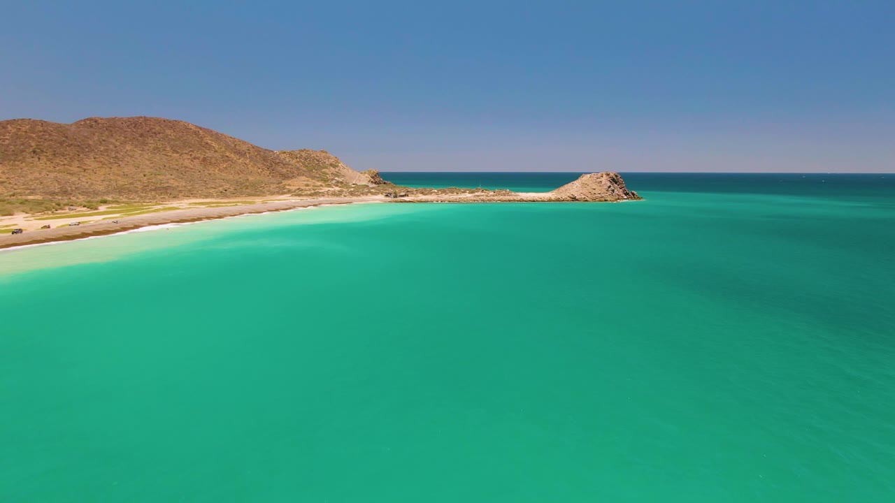 Opening aerial view of Cabo Pulmo with turquoise water in the foreground, sandy beach and dry rocky hills on the left, a small rocky point near the horizon, and no visible people.