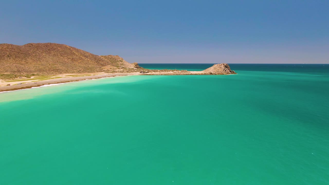 Cabo Pulmo shoreline seen from above, with broad turquoise water across the foreground, pale sand along the left edge, brown rocky hills behind it, and no visible people.