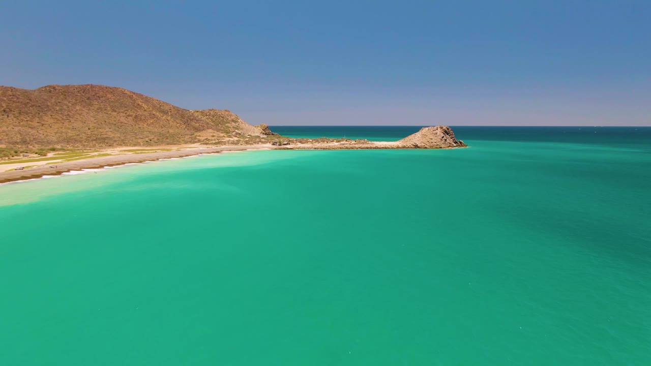 Aerial frame over Cabo Pulmo water, showing the turquoise bay in front, the sandy shoreline and dry hills to the left, a rocky outcrop near the horizon, and no visible people.