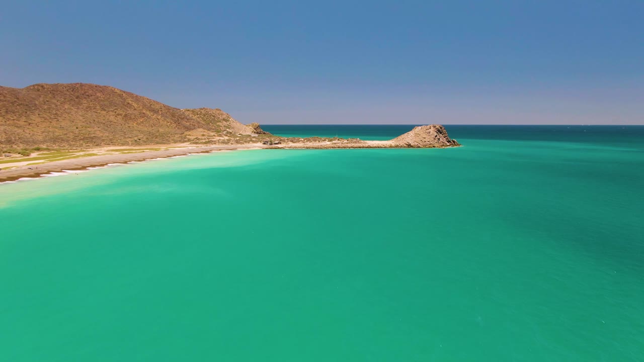 Aerial view of shallow turquoise water beside a sandy beach and rocky hills.