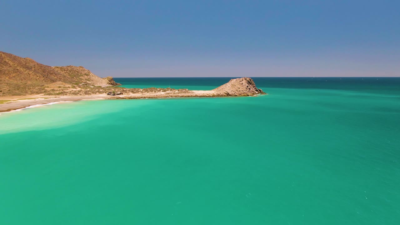 Aerial view of turquoise bay water with sandy beach, rocky hills, and a low outcrop.