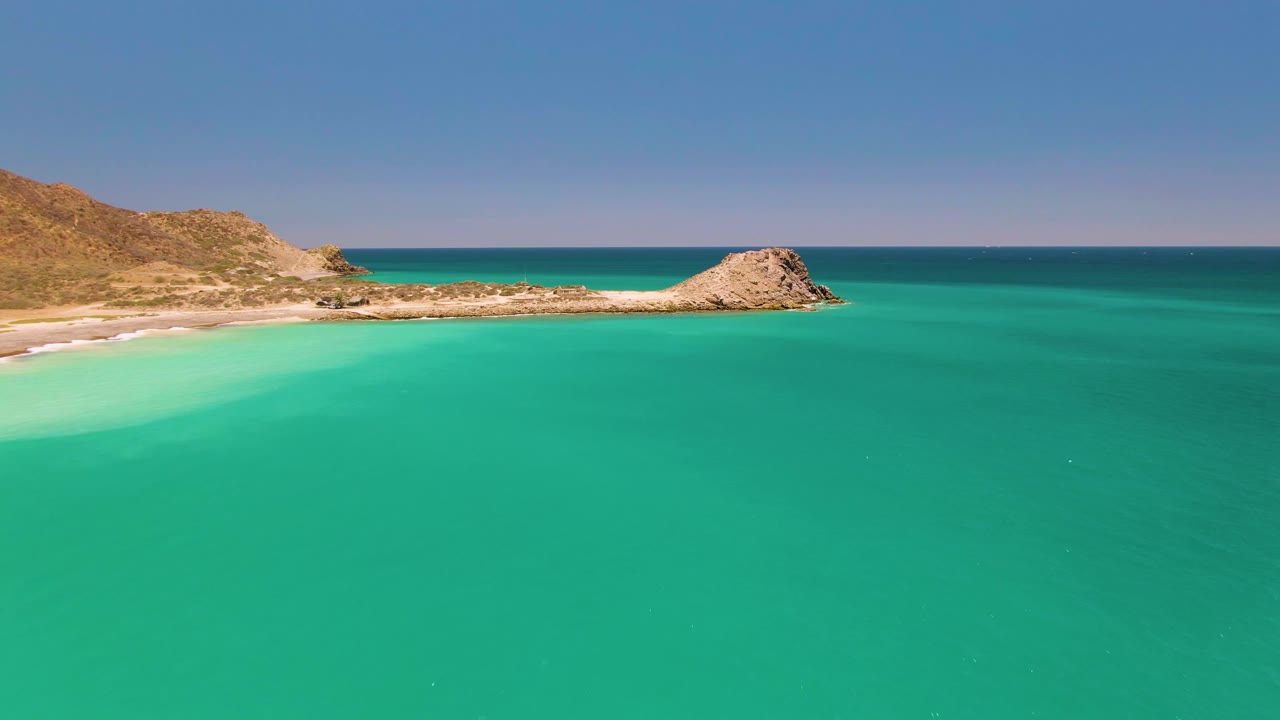 Aerial frame at 0:01 over Cabo Pulmo showing more open turquoise water below the sandy shoreline, with dry hills at left, the rocky outcrop near center, and no visible people.